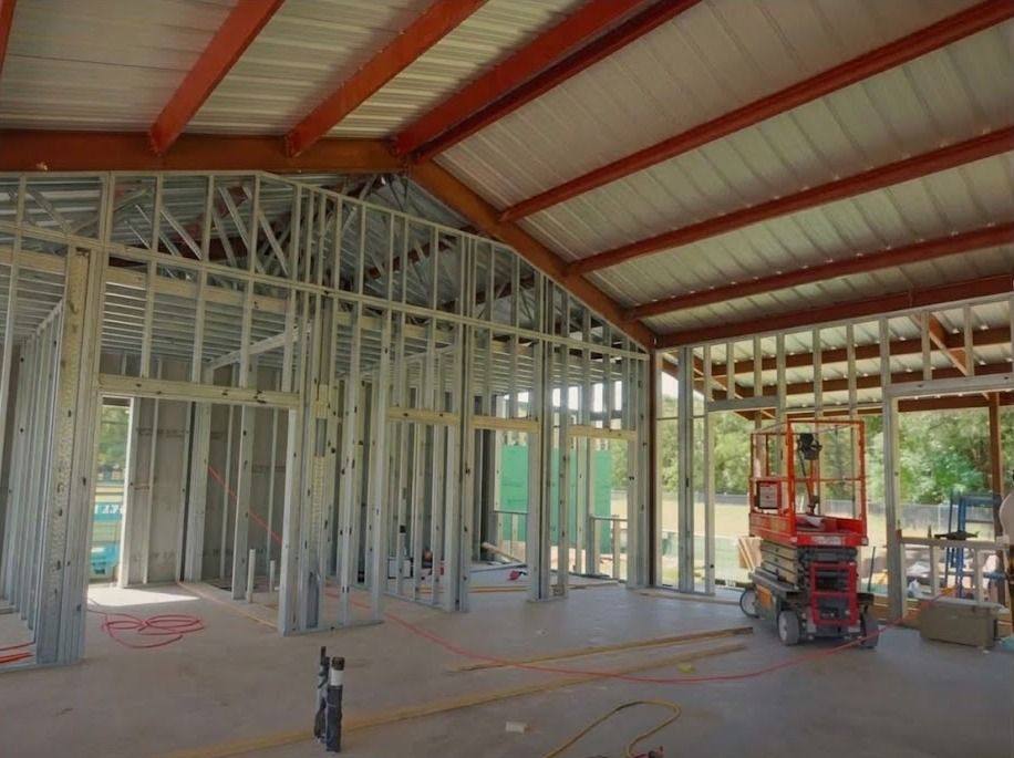 A home mid-construction: red steel frame and metal roof overhead, steel stud interior walls going in on the slab, a scissor lift on site.